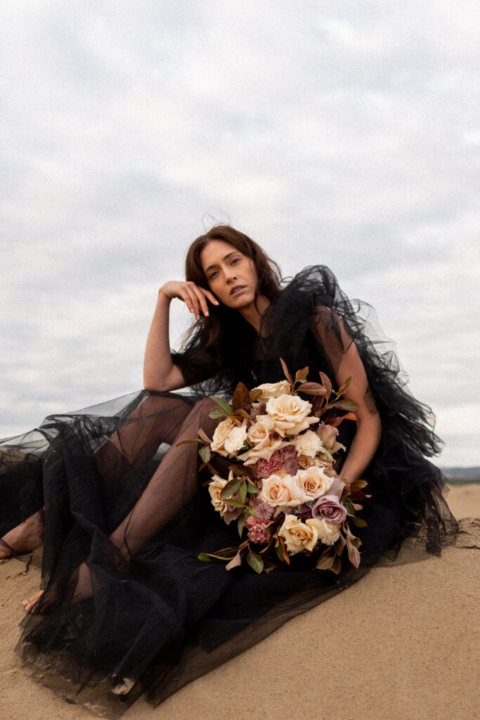 model in a black tulle dress sits in the sand at the little sahara sand dunes in utah