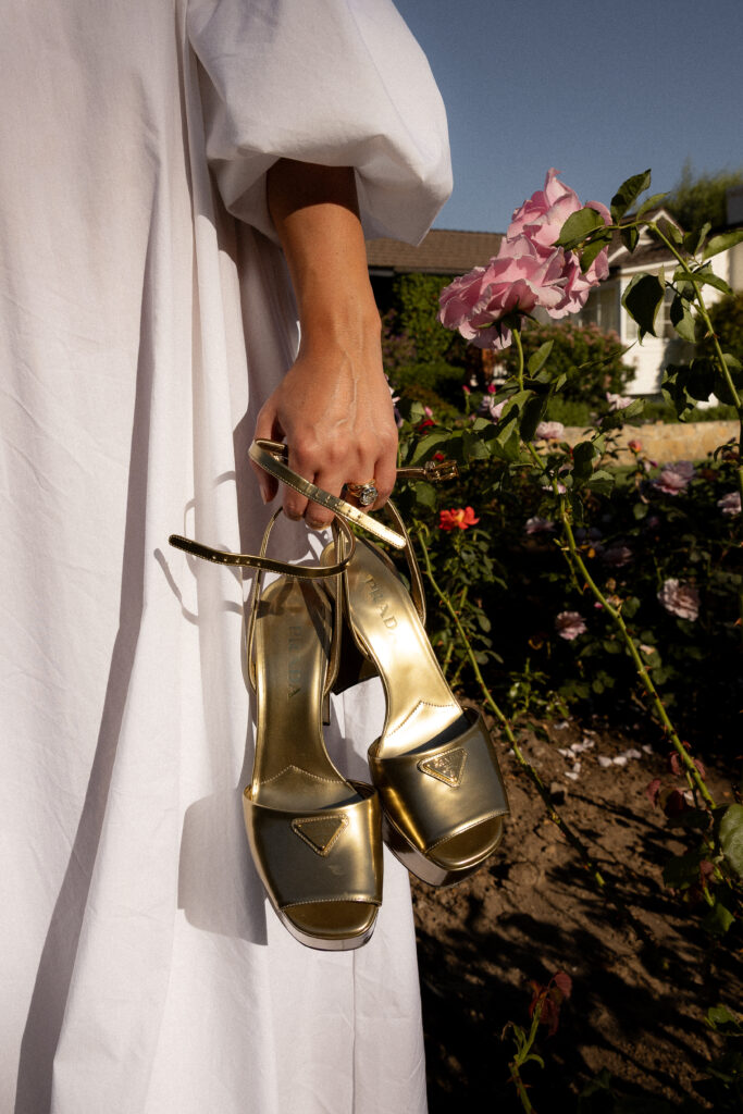 Close-up of a person in a white dress holding gold Prada high-heeled shoes in a flower garden with pink roses in the background.