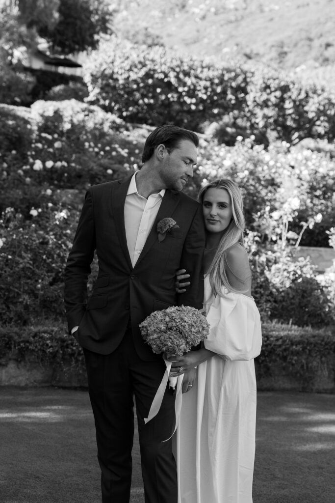 Bride and groom embrace in front of a hillside rose garden at a luxury California estate wedding, with the bride holding a hydrangea bouquet.