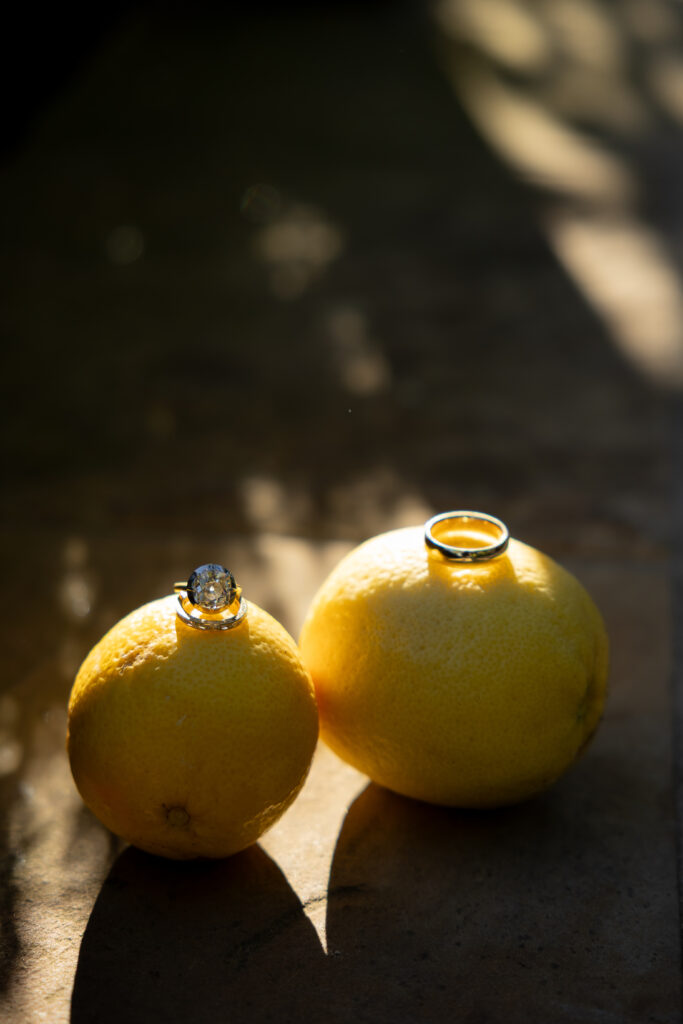 Engagement ring and wedding band placed atop two sun-lit lemons, casting soft shadows on a stone patio.