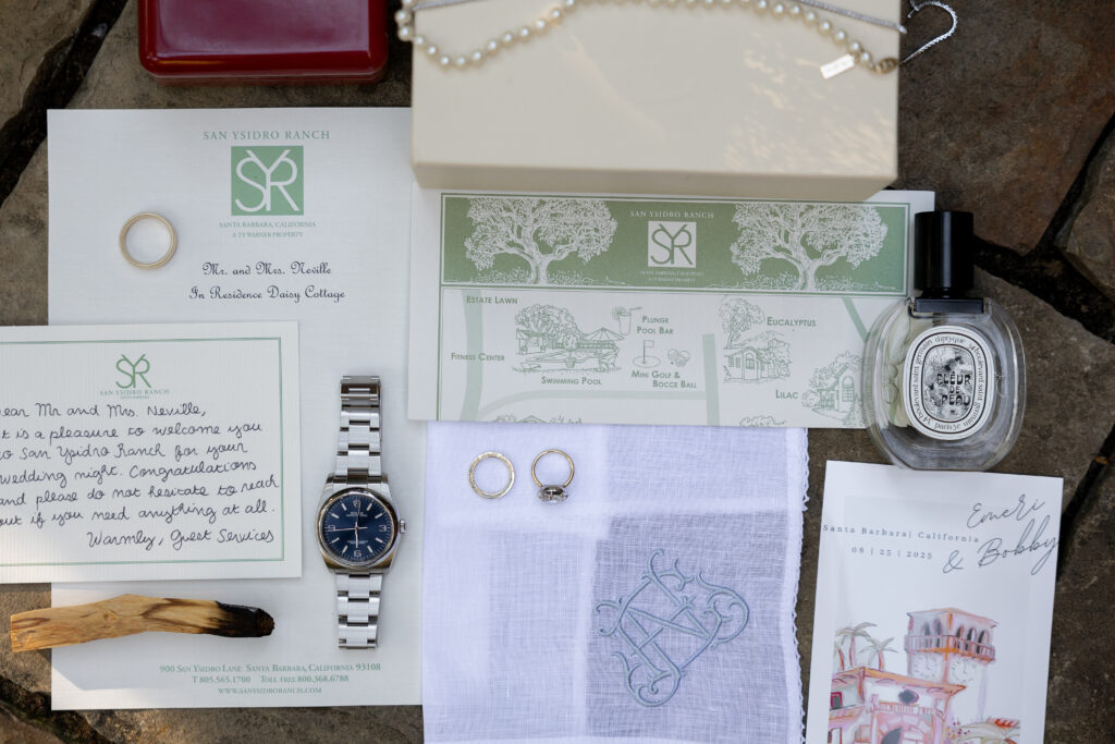 Flat lay of luxury wedding details on a stone surface: a watch, wedding rings, a handwritten note, an invitation suite and perfume bottle, styled together and softly lit.