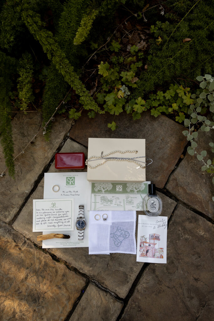 Flat lay of luxury wedding details on a stone surface: a watch, wedding rings, a handwritten note, an invitation suite and perfume bottle, styled together and softly lit.