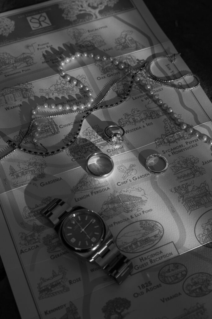 Black and white flat-lay showing a pearl necklace, diamond engagement ring, wedding band and a stainless steel watch arranged on a hand-drawn estate map for a luxury wedding.