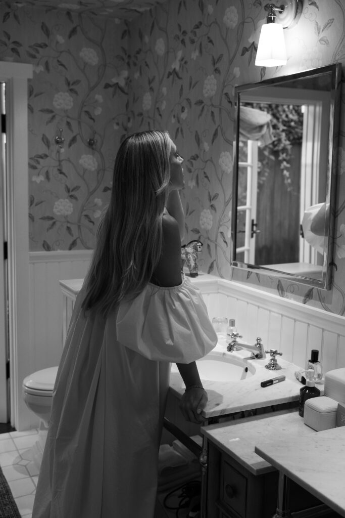Black-and-white photo of a woman in an off-the-shoulder bridal robe, leaning gently on a marble vanity and gazing at her reflection in a softly lit bathroom.