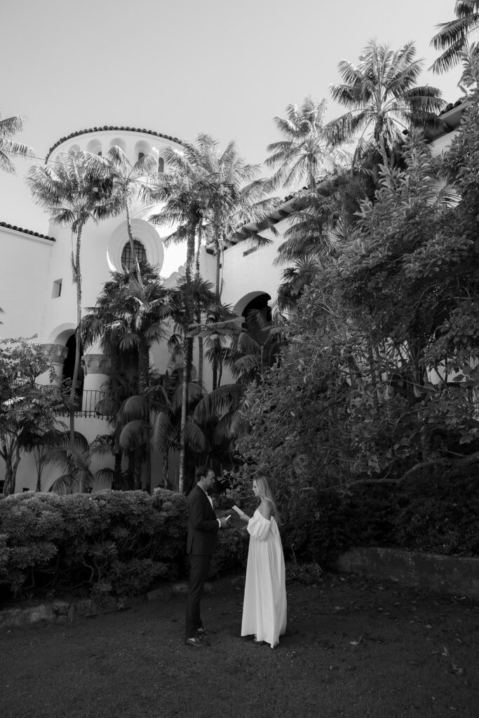 Couple clasp hands mid-vow before towering palm trees in a secluded courthouse courtyard, rendered in elegant black and white.