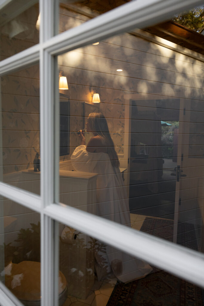Seen through a window reflection, a woman in an off-the-shoulder gown applies lipstick at a bathroom mirror under warm lights.