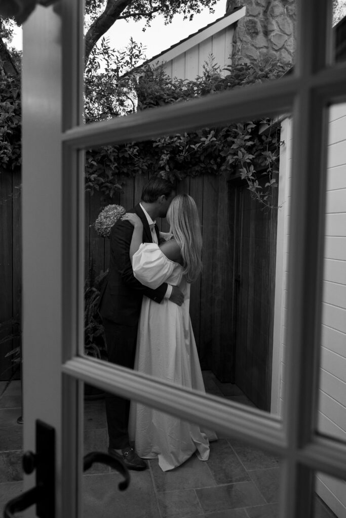Seen through a window, a person in a suit holds a woman in an off-the-shoulder gown as they embrace in a private courtyard surrounded by vines.