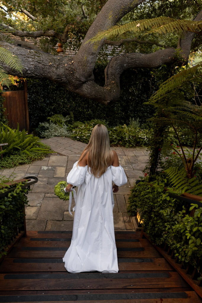 A woman in a flowing off-the-shoulder white wedding gown walks down wooden steps into a garden courtyard, holding a green bouquet, framed by overhanging branches and dense foliage.