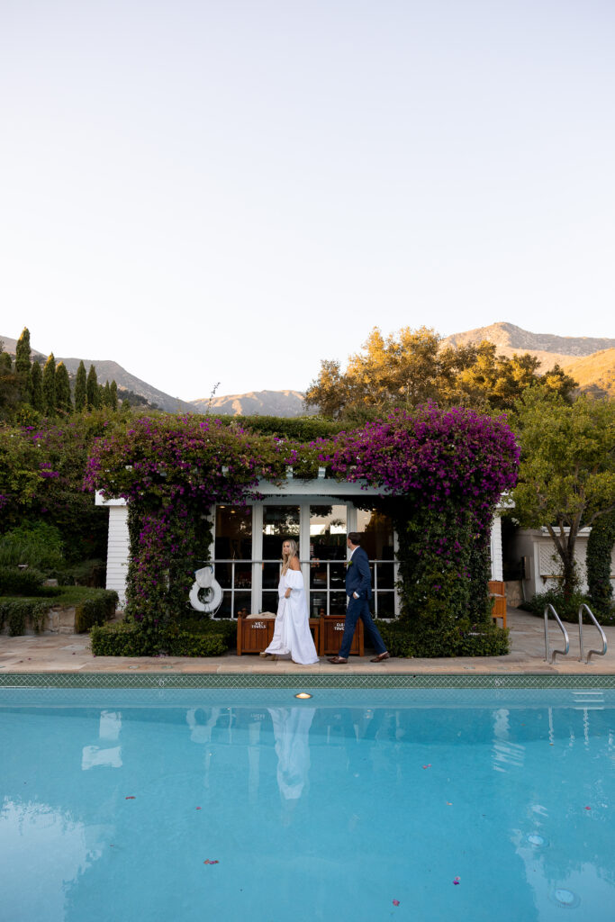 A woman in a flowing white off-the-shoulder gown walks beside a clear blue pool with a man in a suit following her; behind them is a white building draped in vibrant purple bougainvillea, surrounded by trees and mountains.