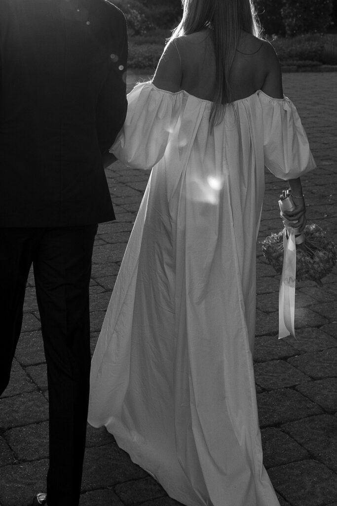 Black-and-white close-up of a couple walking hand-in-hand, seen from behind. The woman wears a flowing off-the-shoulder gown and holds a small bouquet at her side. Sun flares catch the fabric as they walk across stone pavers, creating an elegant, romantic atmosphere