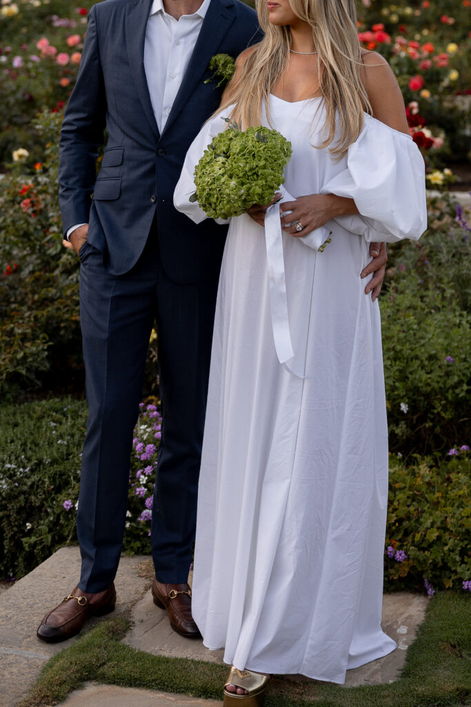 An off-the-shoulder white gown and groom in a navy suit hold hands in a garden wedding portrait with green bouquet, California luxury wedding.