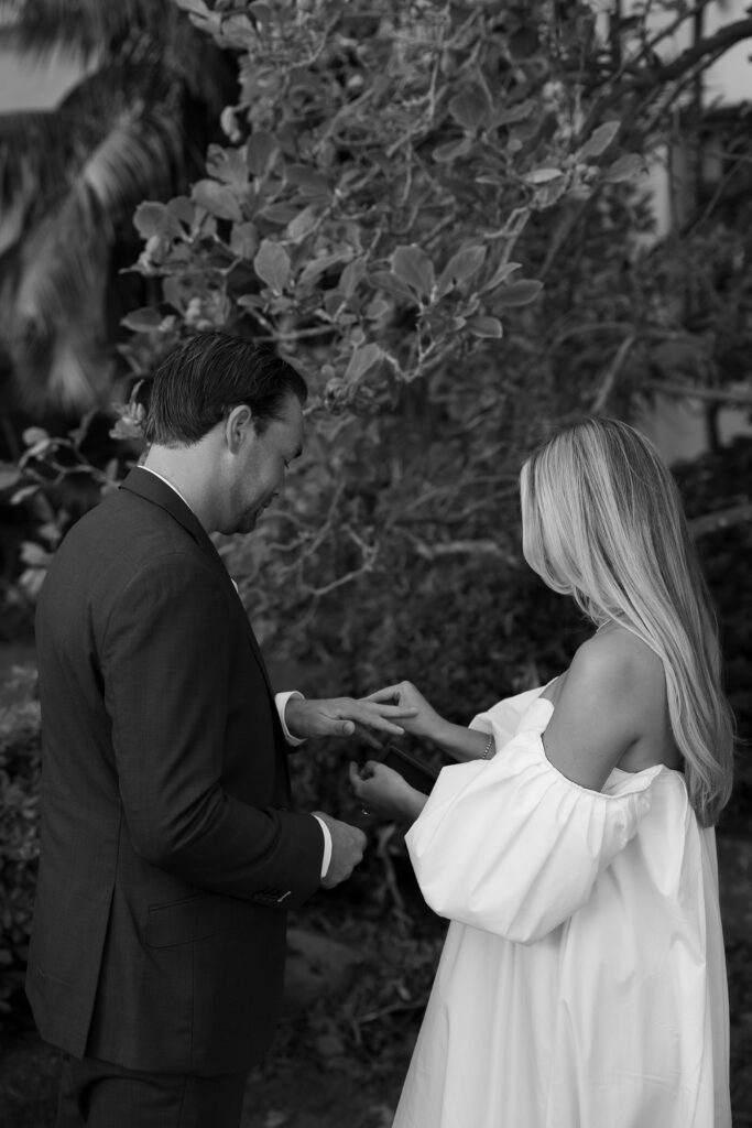 Rings are being placed on the hands of the bride as her hair draped over her shoulders in a black and white photo. 
