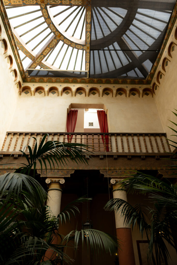 Large glass-roofed terrace ceiling over a courtyard, with giant green palm-leaves softly backlit and a row of tall arched windows along the upper level letting in muted daylight.