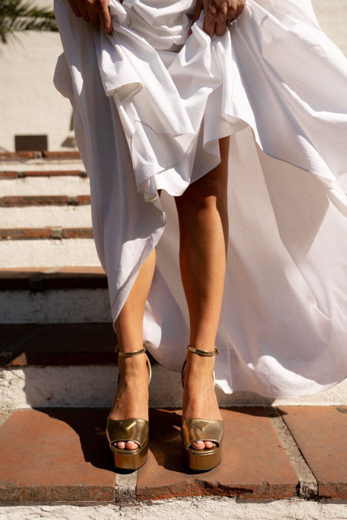 Close-up of bride lifting her wedding dress while walking down terracotta steps, showcasing gold platform heels during a luxury California wedding.