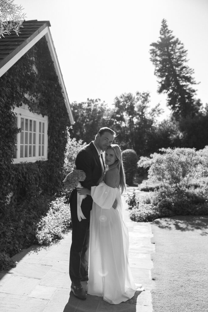 Bride and groom embrace outside an ivy-covered estate during a luxury garden wedding in California, black-and-white portrait.