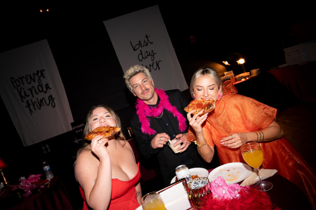 Wedding guests decorated in feathered boa's gather around for a photo while taking bites of take out pizza and a sip of a cocktail!