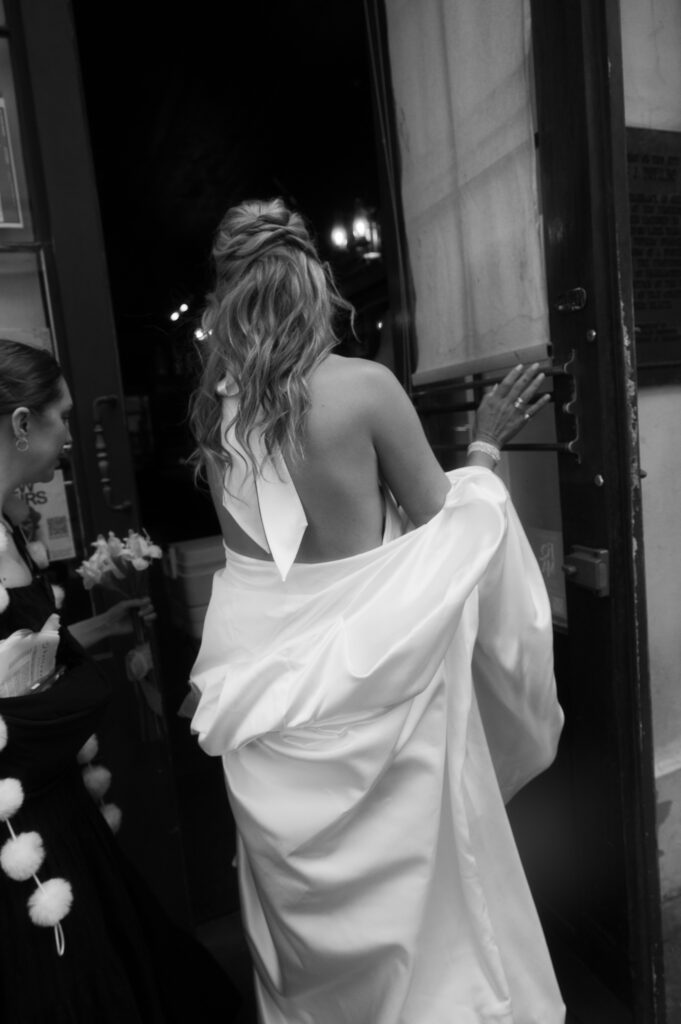 The bride in motion of opening a door shows her hair slightly ruffled as she lightly touches the window pane of the door as her bridesmaid assists her. The black and white aspects of this photo highlight the dramatics of the shadows.