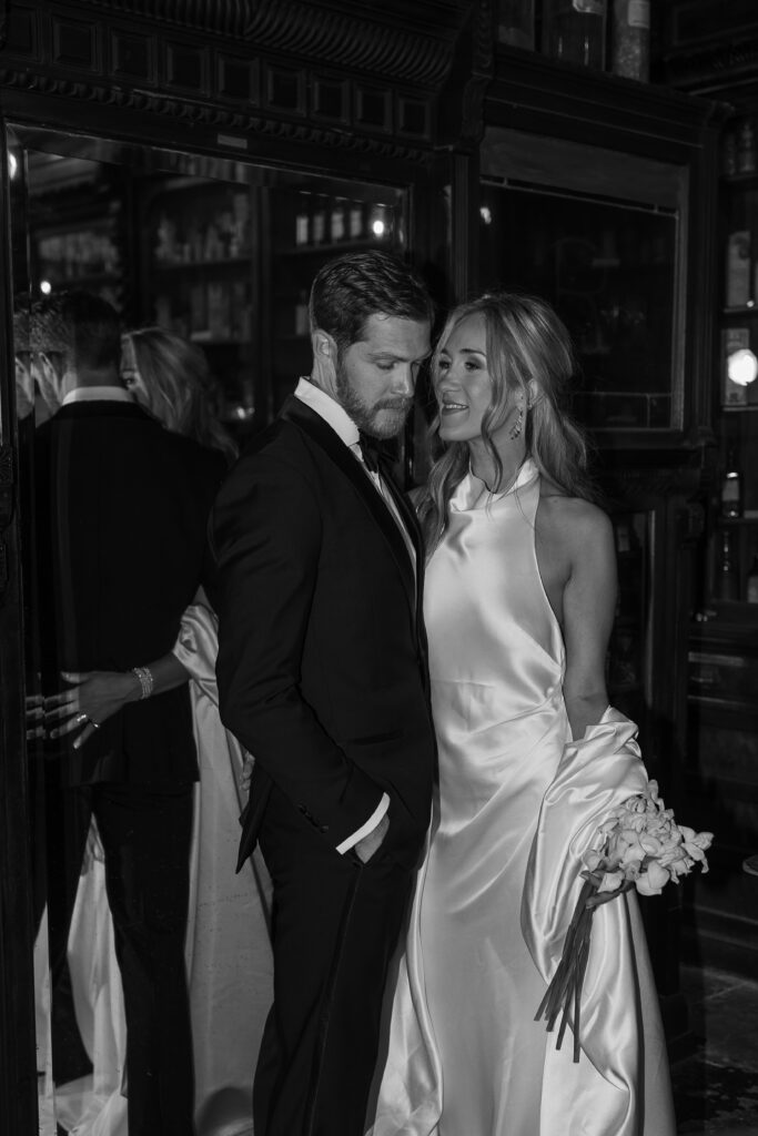 Bride and groom closely embracing one another in a black and white photo, there is a mirror directly behind them which highlights the brides close embrace of the grooms lower back. The bride is wearing a soft satin dress while casually holding up a minimal bouquet. The photo highlights Linden Wilson's Luxury photography.
