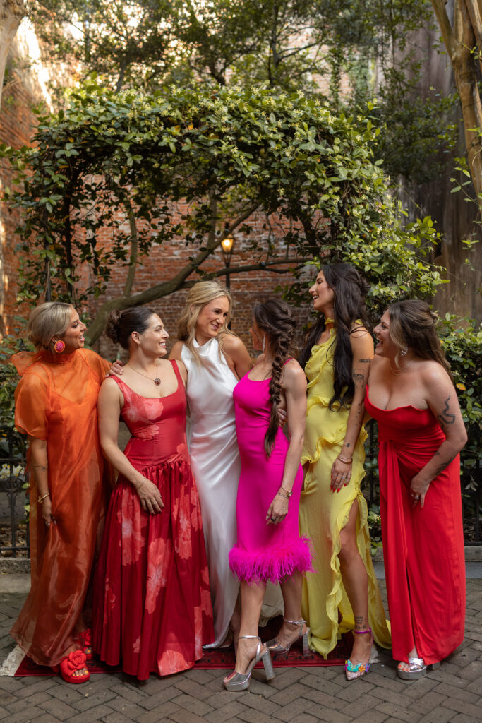 In an outdoor corridor a bride with her 5 bridesmaid are all looking at one another with soft smiles and laughter. The bridesmaids are in very bold colors, textures and patterns. The arbor behind them is full of a heavy greenery, that makes the dress colors pop!