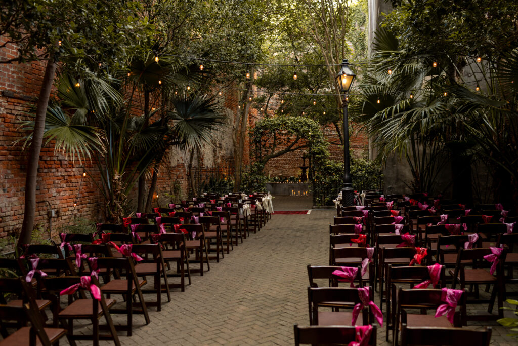 Colorful photo of an empty ceremony space before people fill the chairs that are decorated with red and pink satin ribbon. The ceremony will take place in a concrete and brick corridor under the arbor of deep greenery and string lights hanging from above. A detailed photo for a luxury wedding.