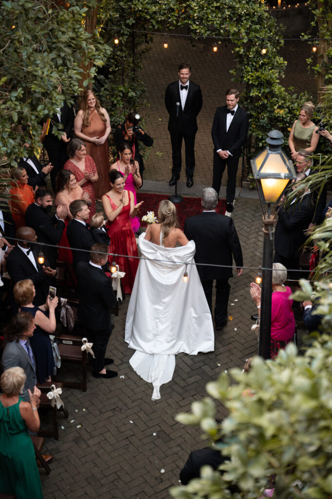 Bride walks down the aisle with her father to meet her groom, her bridesmaids gather around in clapping and cheers while they arrange her dress in a timely manner.