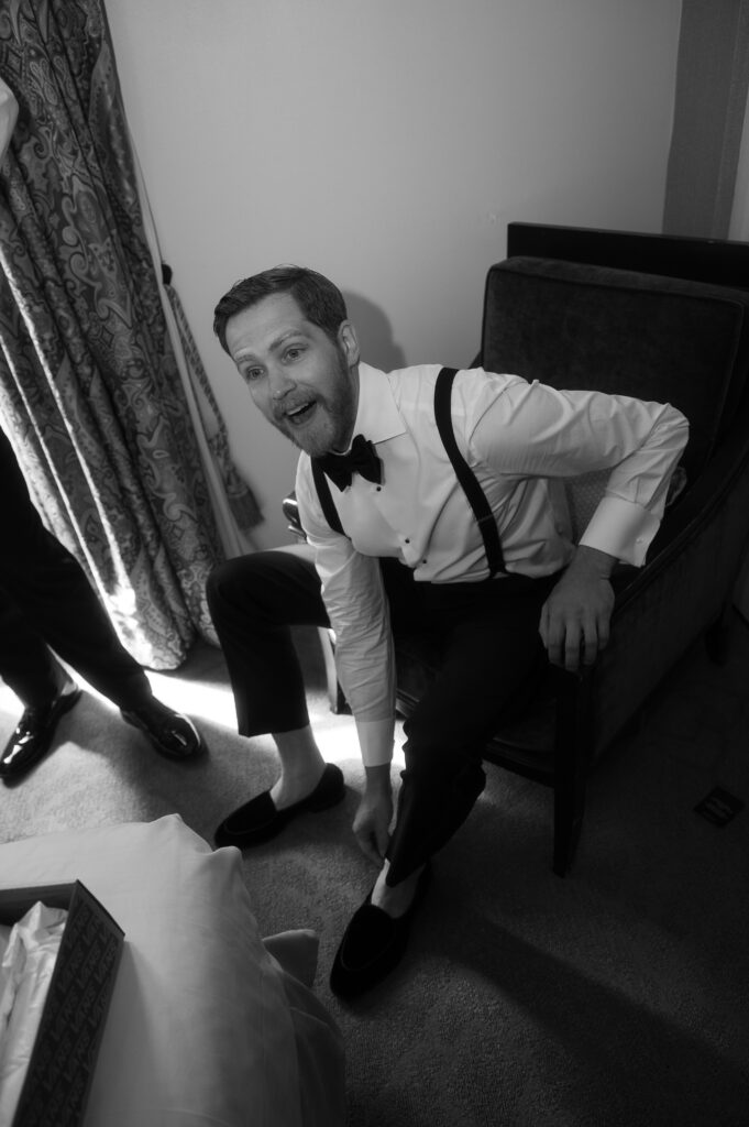 Black and white photo of the groom putting on his black loafers with sharing a deep laugh with his groomsmen.
