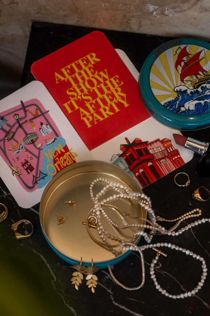 "After the show is the after party" post card shown with lipstick, pearls and the little golden jewelry details, layered outside of a vintage mints can.