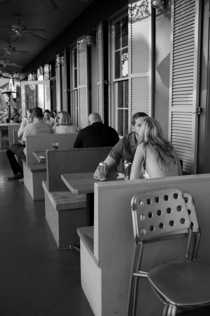 Black & white image of a man and woman in a booth, woman’s back to the viewer, man shifting closer to kiss her.