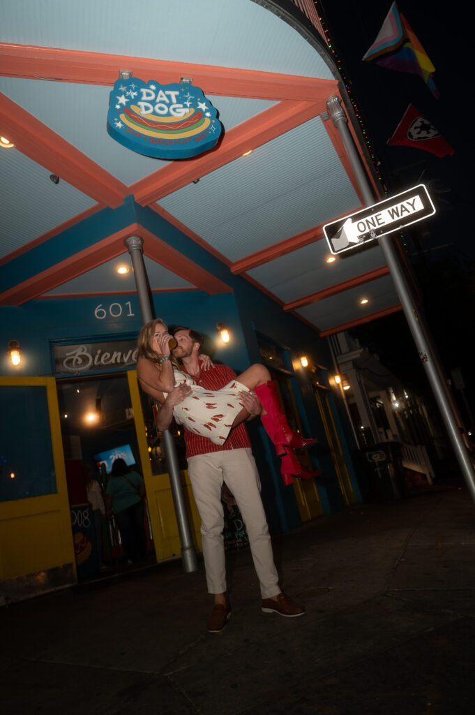 Couple stand in front of a "Dat Dogs" restaurant, in the evening light. The woman is wearing a short white dress with vibrant red chillies and tall red leather boots as she sips a cocktail and the man holds her in his arms while kissing her cheek.