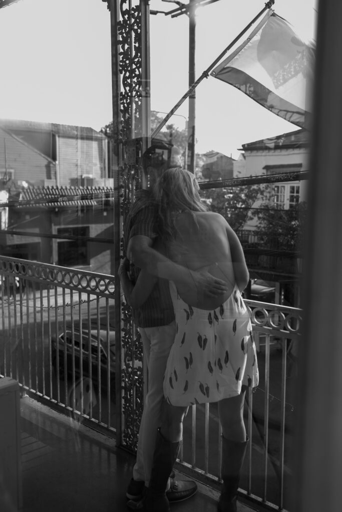 Black and white photo of couple leaning into one another with arms wrapped around the backs of each others. The New Orleans flags are softly flying in the wind as the look over the balcony.