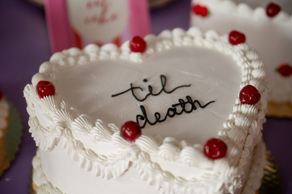Close up photo of white vintage piping on a wedding styled cake. Red candies are on placed as decorations, minimal black piping labeled the cake "til death" on top.