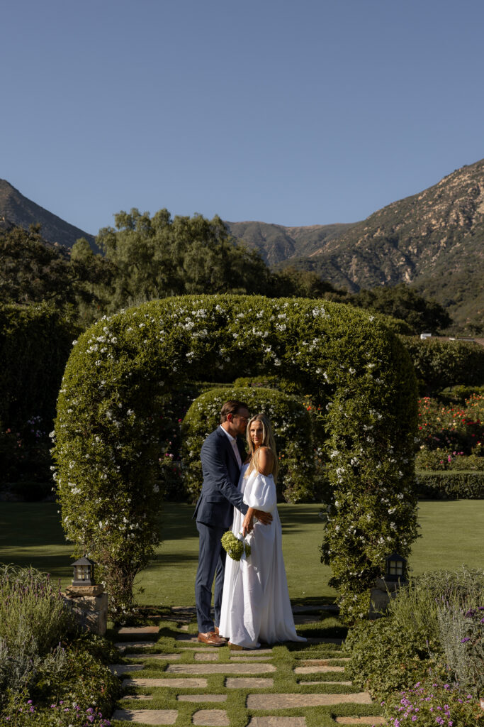 A bride in a luxurious off the shoulder gown glances back at the camera while her groom gently kisses her cheek, standing beneath a well-manicured shrub archway.