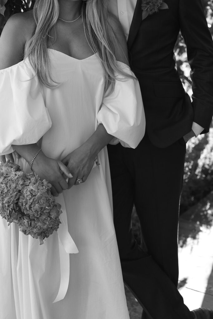 Classic black-and-white portrait of a newlywed couple. The photo focuses on their pose and the elegant details of the bride's gown and groom's suit, while their faces remain unseen, adding a sense of mystery and timeless elegance.